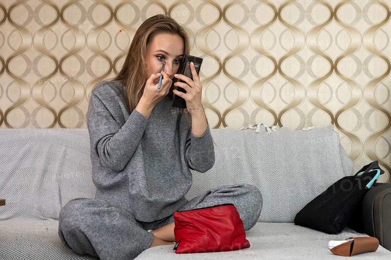 Young Caucasian woman with long brown hair applies makeup in a cozy home setting. She sits on a couch, holding a mirror and a makeup brush