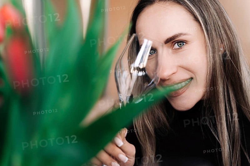 Young long-haired woman smiling while covering one eye with wine glass, A woman smiling while holding a wine glass with green plants near, creating a casual and joyful atmosphere