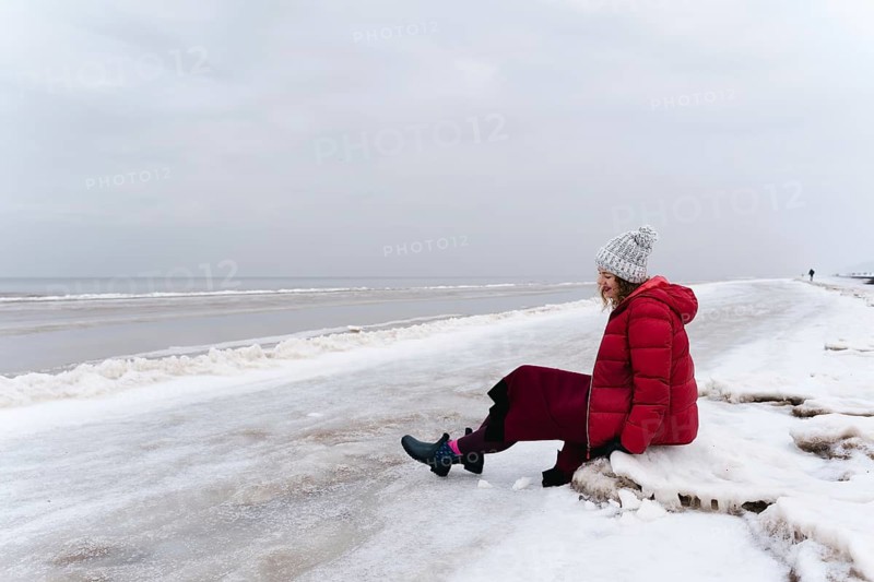 A cheerful woman in a red jacket and a warm dress sits on the shore of the winter sea, A woman in a red coat and gray hat sits on a snowy beach, looking out at the ocean under a cloudy sky, Jurmala, Latvia
