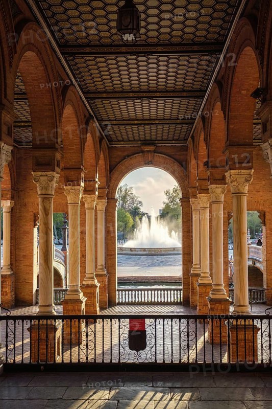 Framed view of Plaza de Espana Public Square in Seville, Spain, showing arched gallery, central fountain, brick arcade, and marble column