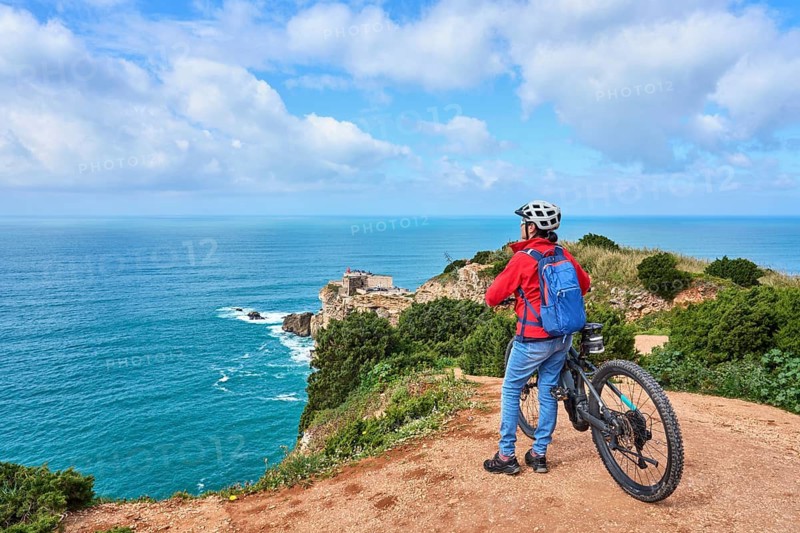 Attractive senior woman riding her electric mountain bike on the rocky cliffs of Nazare. western atlantic coast, Portugal