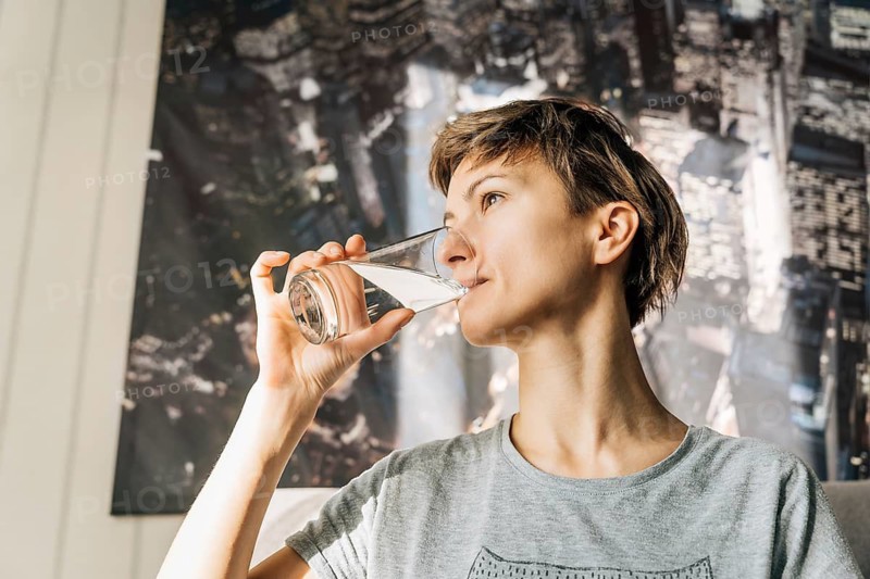 A young woman with short hair drinks water from a glass, Woman in a gray shirt drinks water from a glass, sitting indoors with sunlight streaming in
