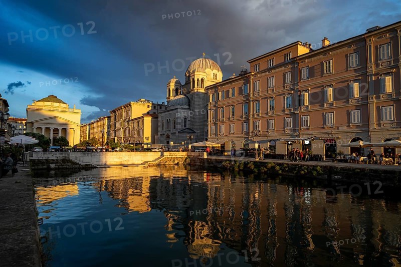 Grand Canal with restaurants, Sant'Antonio Taumaturgo church and Serbian Orthodox church Santissima Trinità e di San Spiridione, reflection in the canal, in the evening light at sunset, dramatic clouds and atmospheric lighting, Borgo Teresiano, Trieste, Friuli-Venezia Giulia region, Italy