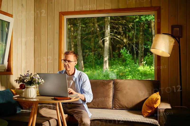 Middle-aged man is sitting on sofa in wooden cabin, focused on his laptop. He looks at latest quotes for stocks he has invested in. Outside window green trees are visible under sunlight