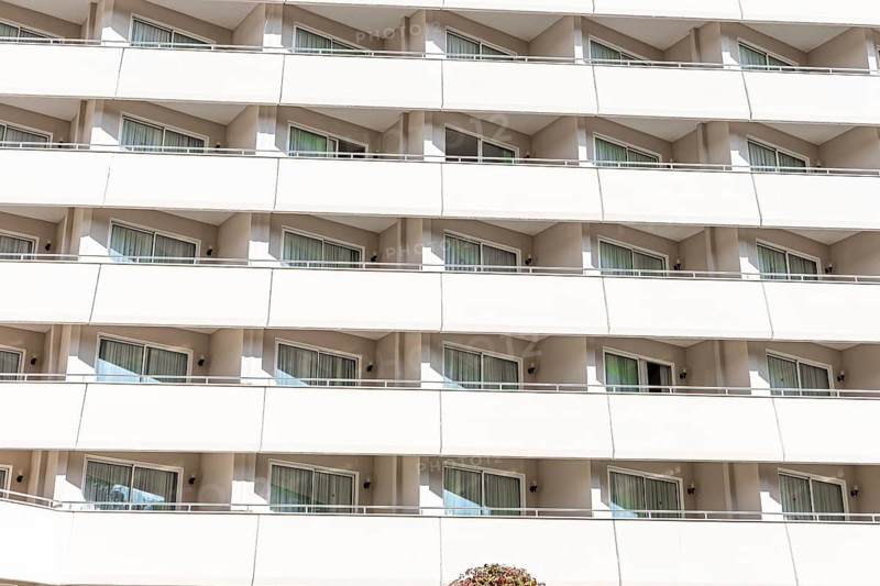 A modern hotel building with multiple balconies and large windows. The structure features a clean, white facade and is surrounded by greenery