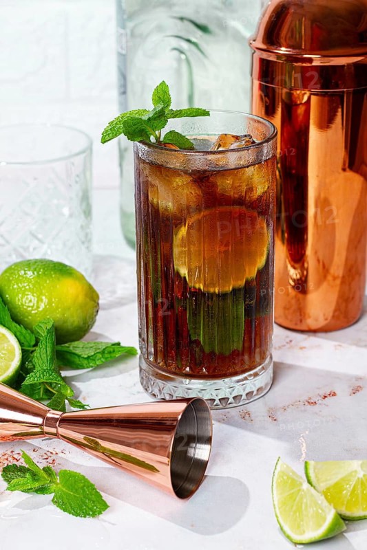 A tall glass filled with a dark soda drink shows slices of lime and fresh mint on a white table. A copper shaker sits in the background with other glassware nearby