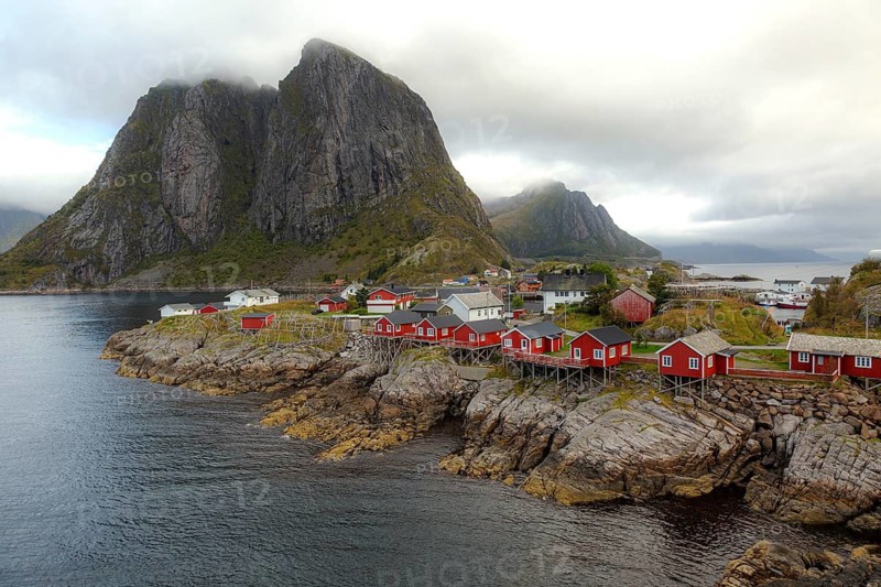 Picturesque coastal landscape of Hamnøy with traditional red fishing huts, rocky coast and the imposing Festhæltinden under dramatic cloudy skies, Hamnøy, Lofoten, Norway