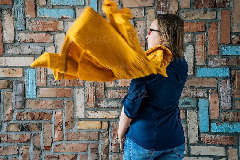 Rear view of a 40-year-old woman in glasses with a flying yellow jacket against a colorful brick wall, A woman in a blue shirt and yellow scarf stands against a textured brick wall, with the scarf in motion