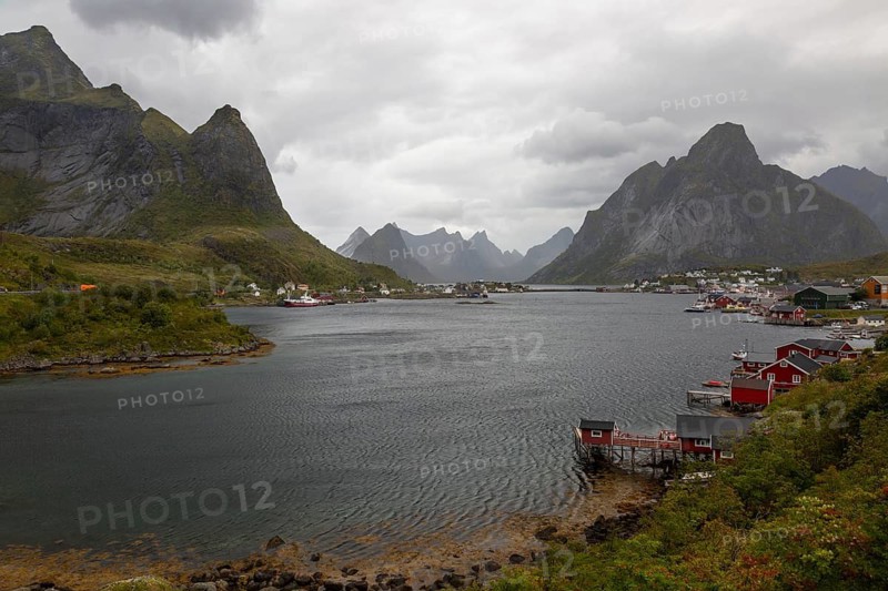 Picturesque bay of Reinefjorden surrounded by the Reinebringen and Olstinden mountains with red Rorbuer huts on the shore under dark cloudy skies, Reine, Lofoten, Norway