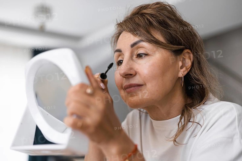 A mature woman is enjoying her beauty routine as she applies mascara. She smiles at her reflection in a handheld mirror, showcasing a moment of confidence and self-care in a cozy setting