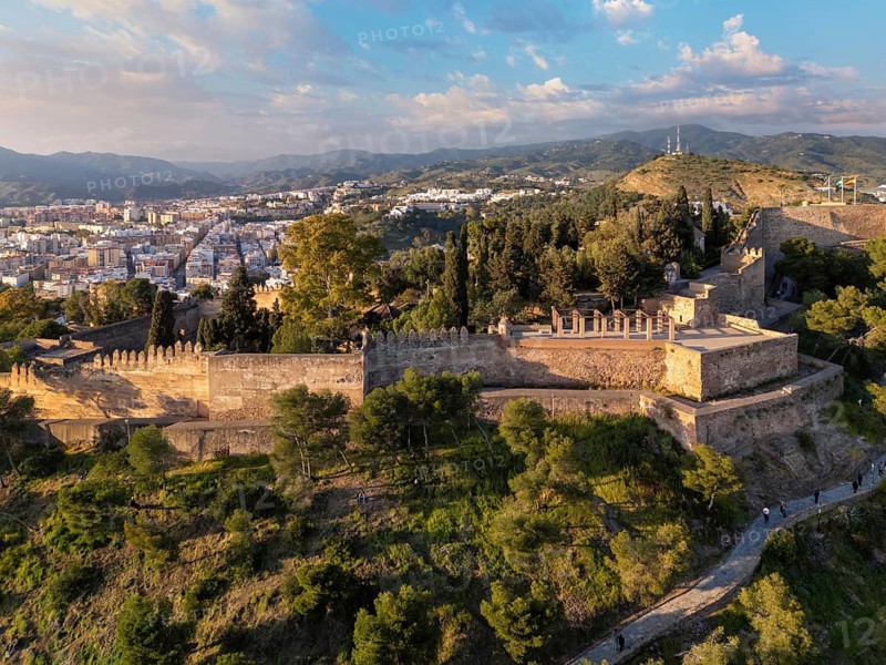 Close aerial view of Gibralfaro Castle in Malaga, Spain, showing sunlit battlements, defensive walls, wooded slopes and the city beneath a cloud-filled sky, featuring castle walls