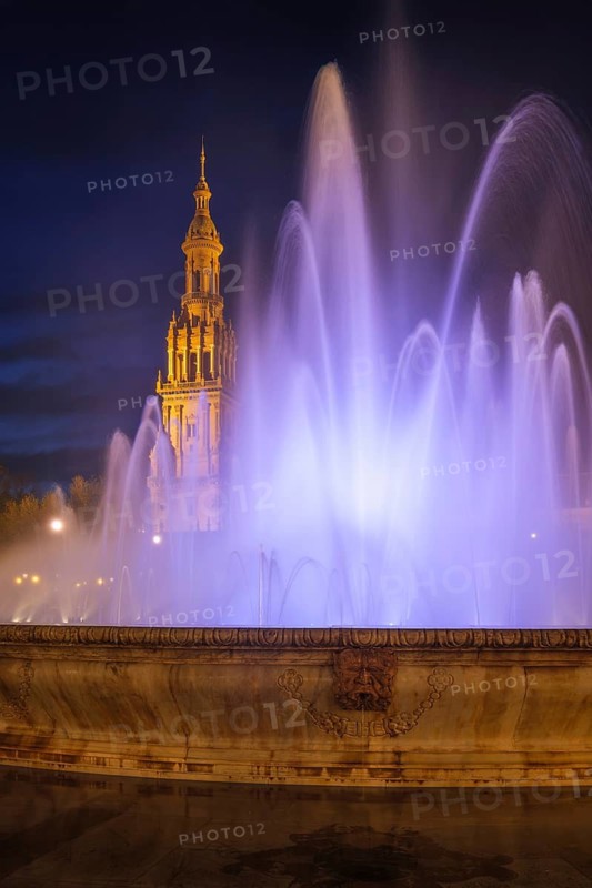 White and lavender fountain jets frame the illuminated North Tower of Plaza de Espana in a vertical night composition in Seville, Spain, featuring water fountain and colored lighting