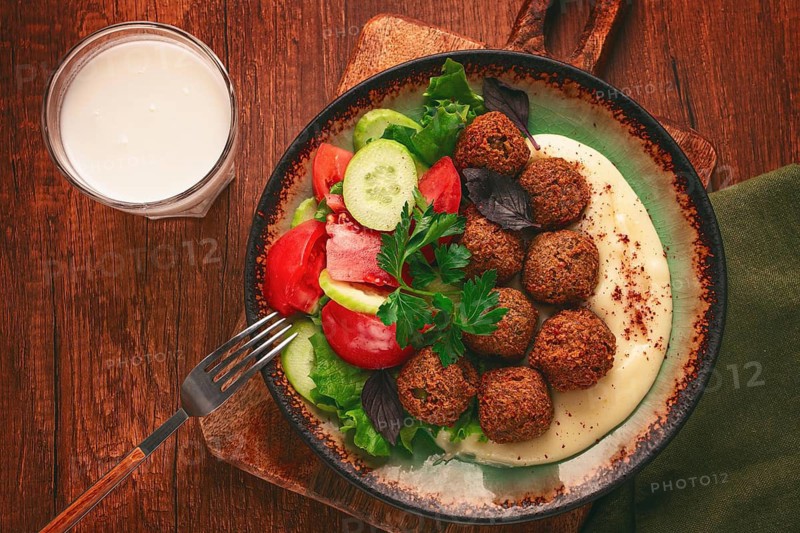 A plate holds falafel balls, tahini, salad with tomatoes and cucumbers, and a drink. The setting is warm and inviting, perfect for a midday meal with fresh ingredients