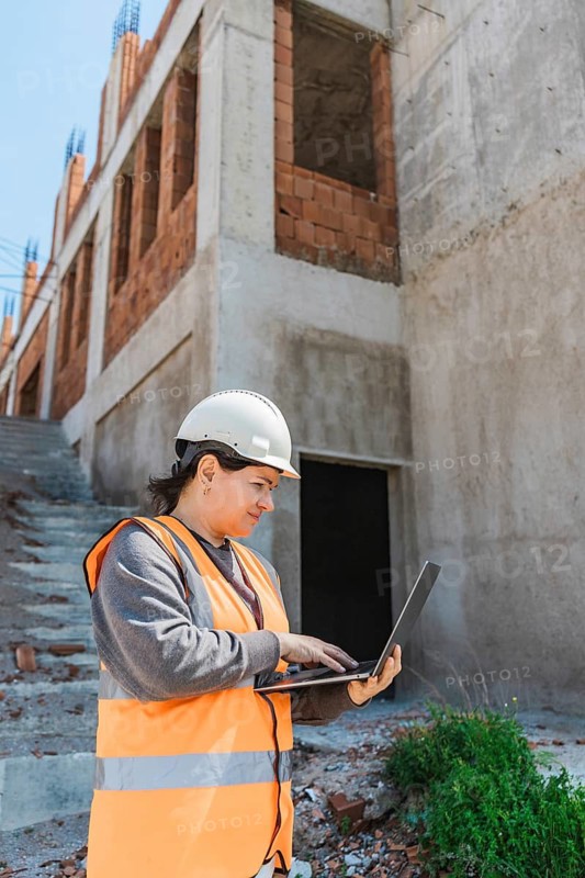 Woman in a hard hat and safety vest uses a laptop on a construction site. The background features unfinished buildings and stairs