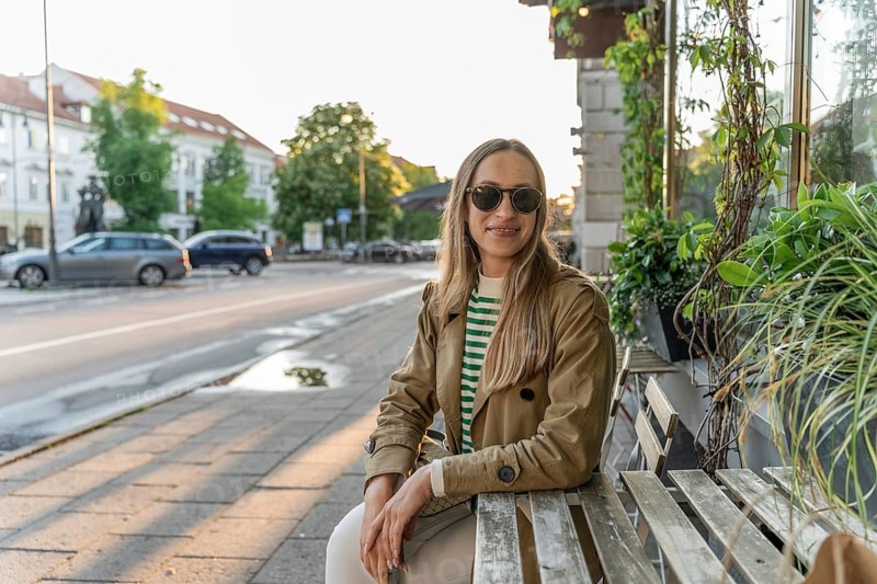A woman in sunglasses sits at the table of an outdoor cafe, A woman wearing sunglasses sits on a bench beside a street with plants and sunlight in an urban setting, Vilnius, Lithuania