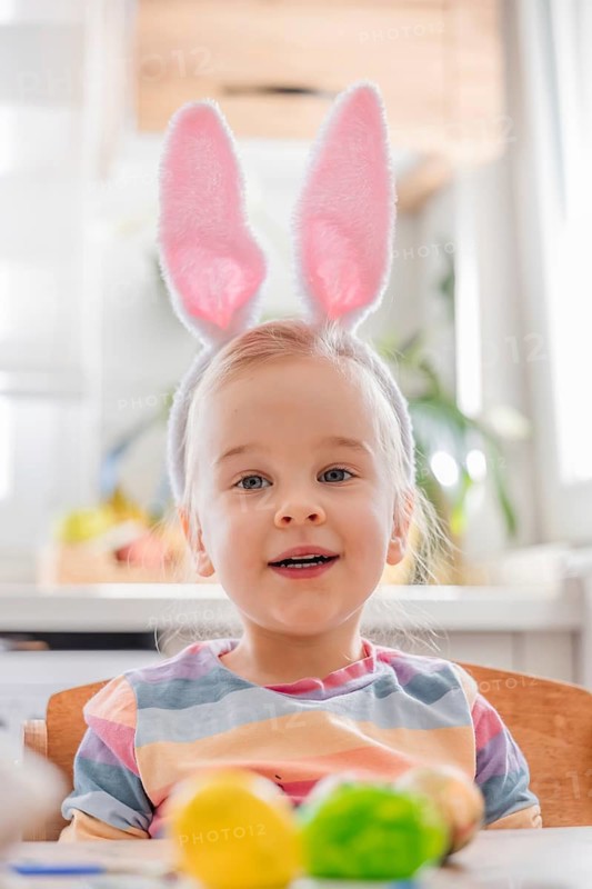 A child wearing bunny ears sits at a table decorated for Easter. Colorful eggs are placed on the table. The room has bright light and festive items. The child looks cheerful and excited
