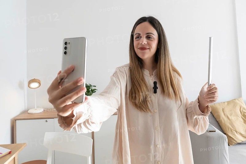 Close up of smiling woman holding smartphone and LED light panel, recording video content with lavalier microphone clipped to her blouse