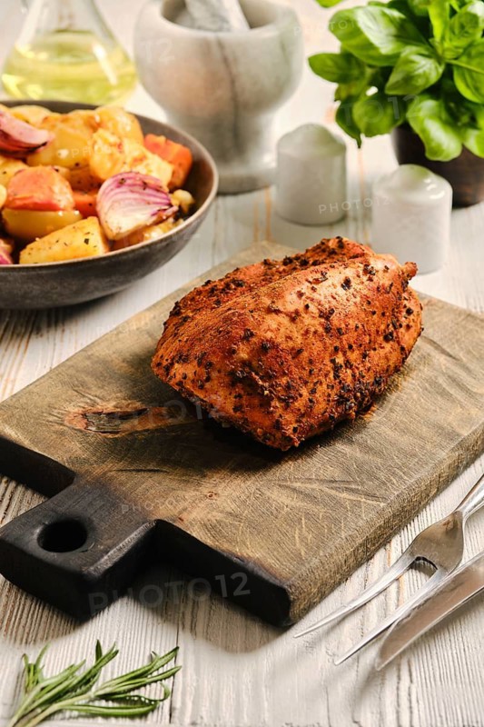 Roasted turkey breast on wooden cutting board in kitchen. Nearby bowl with vegetables. Oil and herbs are visible in background