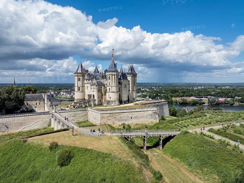 Aerial view of the historic Château de Saumur with its distinctive round corner towers and bright facades, surrounded by well-kept green spaces under a slightly cloudy summer sky, with the town of Saumur on the Loire River, Maine-et-Loire, Pays de la Loire, France