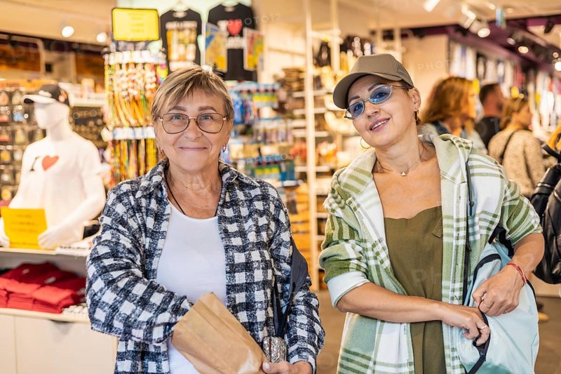 Two women tourists in a souvenir shop. One is a senior Caucasian woman with glasses and short hair, the other is a middle-aged woman wearing a cap and sunglasses