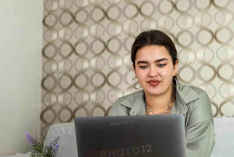 A young Hispanic woman with long brown hair works on a laptop at a wooden table in a cozy living room. A small plant is on the table