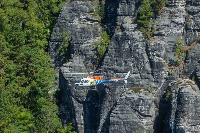 The clean-up work around the health resort of Rathen began immediately after the storm event at the end of July. In the area of the rock stage, fallen trees must be flown out of the spectator area and from the roof of the main building. In addition, trees must be secured and removed from the adjacent rocks that are at acute risk of falling. Areas around the blackbird lake currently resemble a game of Mikado with trunks lying on top of each other. Preparations for the helicopter operation have been ongoing for just over a week. Foresters from Sachsenforst and contracted companies work their way through the storm wood with chainsaws, winches help to loosen and pull apart the logs that are wedged into each other. Tree climbers prepare to fly out trees at the rock stage, Rathen, Saxony, Germany