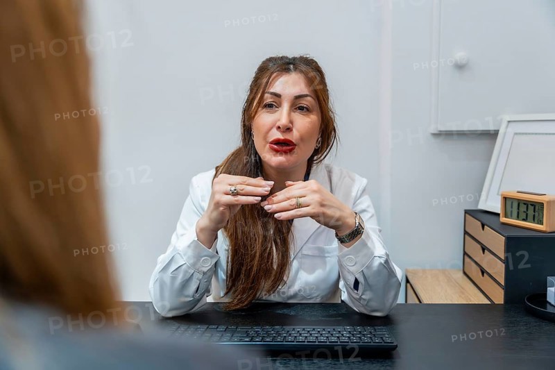 Female doctor in a white lab coat sitting at a desk, actively gesticulating while explaining a treatment plan to an unseen patient during an in person consultation in an aesthetic clinic