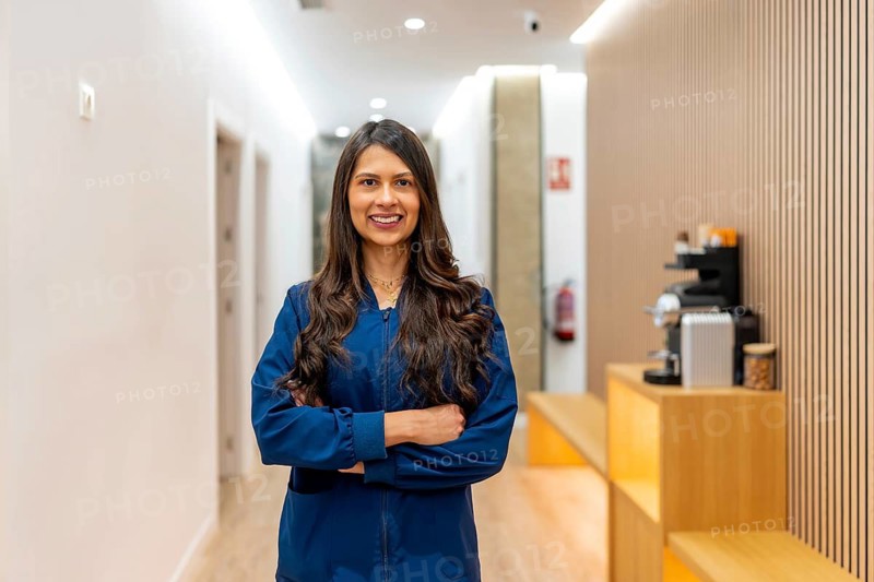Professional woman smiling, wearing a blue uniform, and standing with crossed arms in a brightly lit hallway of an aesthetic clinic or medical spa, ready to welcome clients
