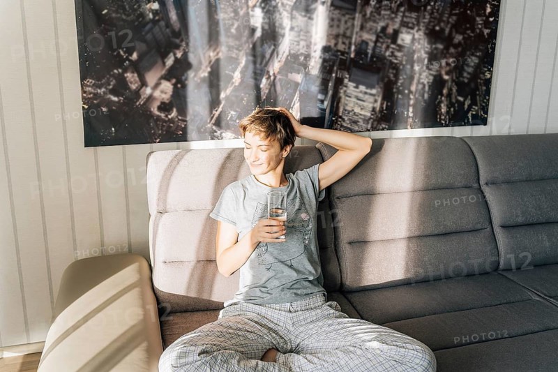 A satisfied young woman with her eyes closed holds a glass of water and smiles, A young man in pajamas sits on a couch in sunlight, holding a glass and appearing relaxed