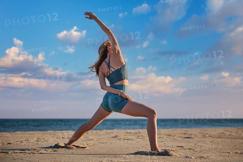 On beach sand, a woman holds a half-bound Viparita Virabhadrasana yoga variation, Reverse Warrior, with one arm overhead and the other behind her back