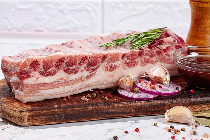 A piece of raw pork rests on a wooden board in a kitchen. Chopped onions, garlic, and a sauce are placed nearby. Spices and bay leaves are scattered on the board