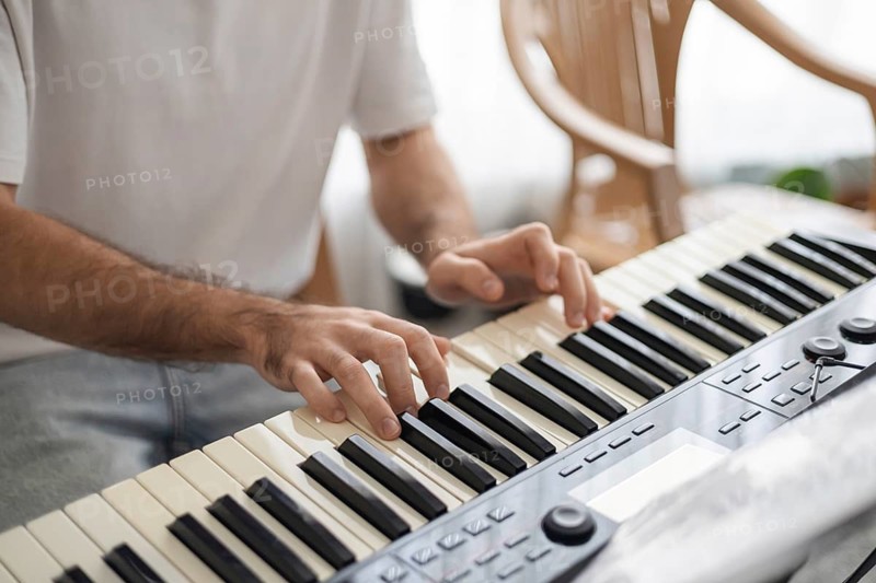 A young man with dark hair plays a keyboard at home. The setting is cozy, with wooden chairs and natural light, creating a warm atmosphere for family music