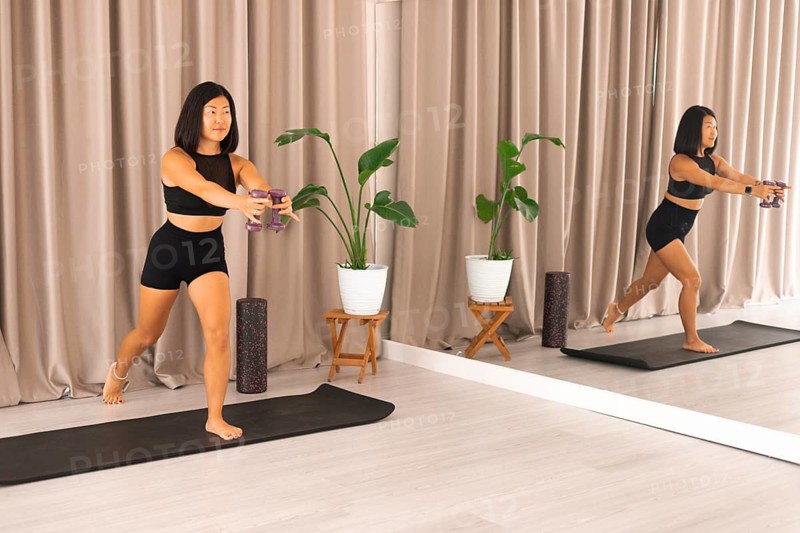 A woman balances while lunging with dumbbells in a room featuring plants and a mirror