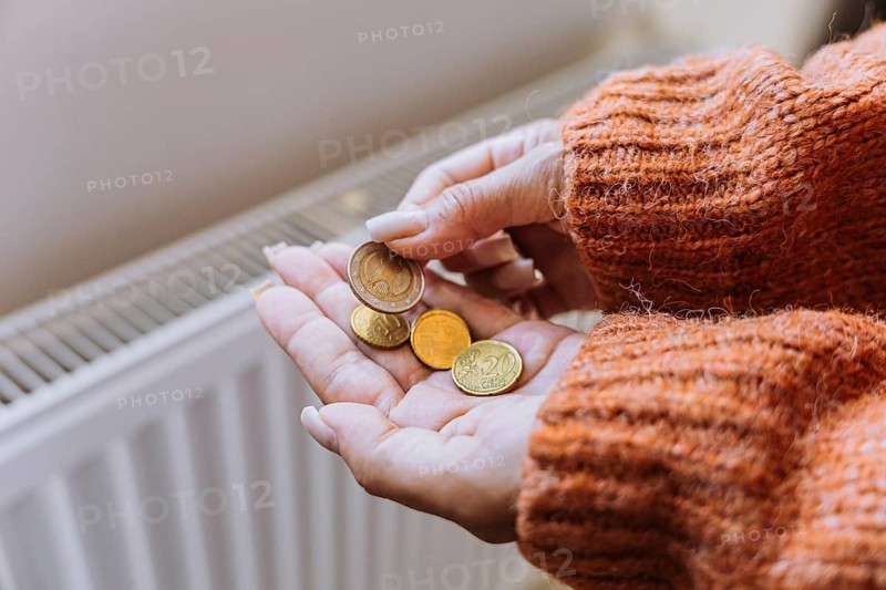 Woman holding euro coins near home radiator during winter heating season in Europe. Concept of rising heating expenses, energy crisis, inflation, difficult household choices, reduced ind.