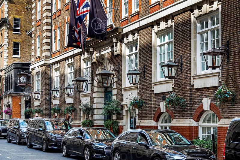 Black cars are parked along the brick and stone facade of the Great Scotland Yard Hotel in Whitehall, its lamps and hanging baskets lining a narrow historic street once home to the Metropolitan Police detective department, London, England, United Kingdom