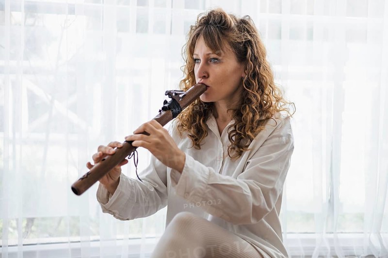 A woman plays a wooden flute by a window, creating a serene musical atmosphere