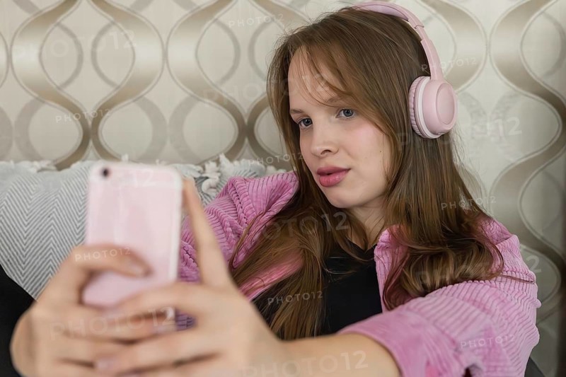 Young person sits on couch wearing headphones while using smartphone. The background shows a decorative wall and soft pillows. Sunlight fills the room in the afternoon