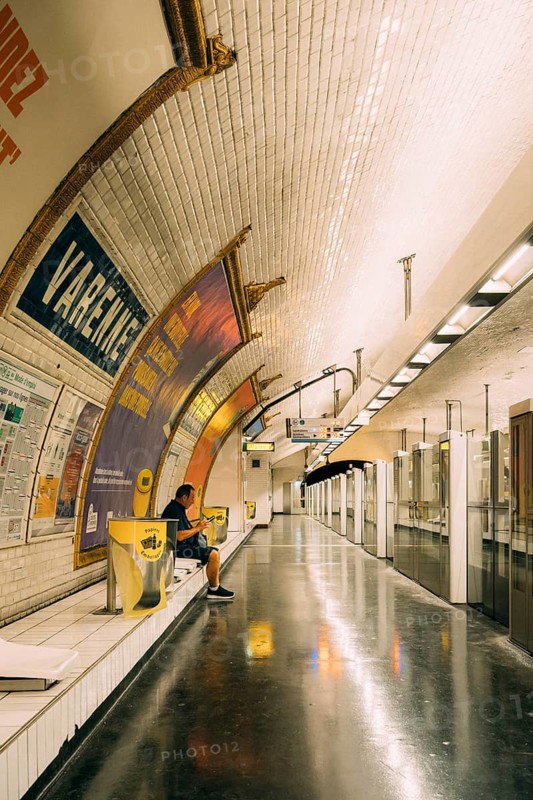Underground platform of a subway station with a vaulted, white-tiled ceiling, large advertising posters on the left wall and a person waiting on a bench, on the right, glass platform doors delimit the track area with reflecting, dark flooring. Paris, Paris Metro, Ile-de-France, France