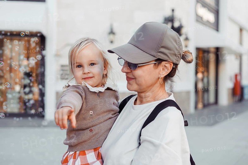 A little girl playfully points towards bubbles while being held by her smiling grandmother. They are enjoying a sunny afternoon in a lively plaza, surrounded by shops and a cheerful atmosphere
