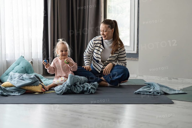 A young Caucasian woman with long brown hair sits on a mat with a toddler girl. The girl has blonde hair and is playing with toys. They are surrounded by blankets in a cozy home setting