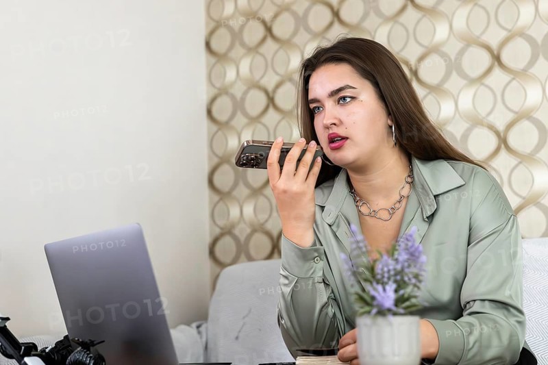 Young Hispanic woman with long dark hair speaking on a smartphone. She wears a light green blouse and a silver necklace. A laptop is visible in the background