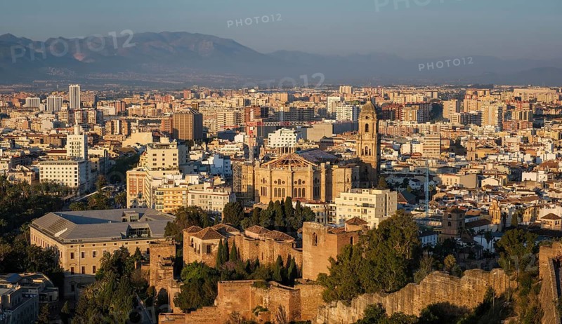 Panoramic elevated view of Malaga, Spain, with the Cathedral and Alcazaba dominating the sunlit historic center while urban districts spread toward layered mountains, featuring cathedral tower