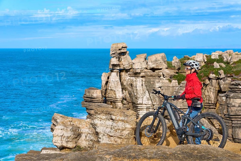 Senior woman riding her electric mountain bike on the rocky cliffs of Peniche at the western atlantic coast of Portugal