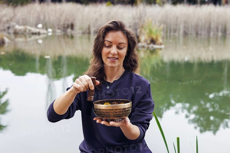A young Caucasian woman with brown hair sits by a serene lake, holding a wooden bowl filled with colorful ingredients. Green reeds surround the water
