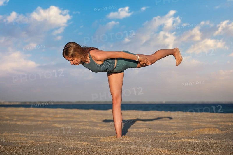 In a standing balance on beach sand, a woman holds a Virabhadrasana 3 yoga variation, Warrior 3 Pose, with arms reaching backward and a slightly bent raised knee