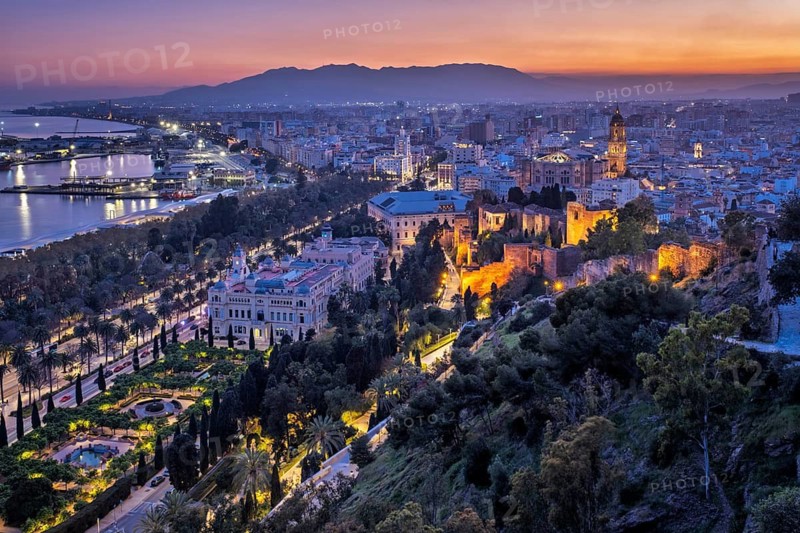 Wide twilight panorama of Malaga, Spain, with illuminated Cathedral and Alcazaba walls above the port, and a dark mountain ridge at sunset, featuring elevated view