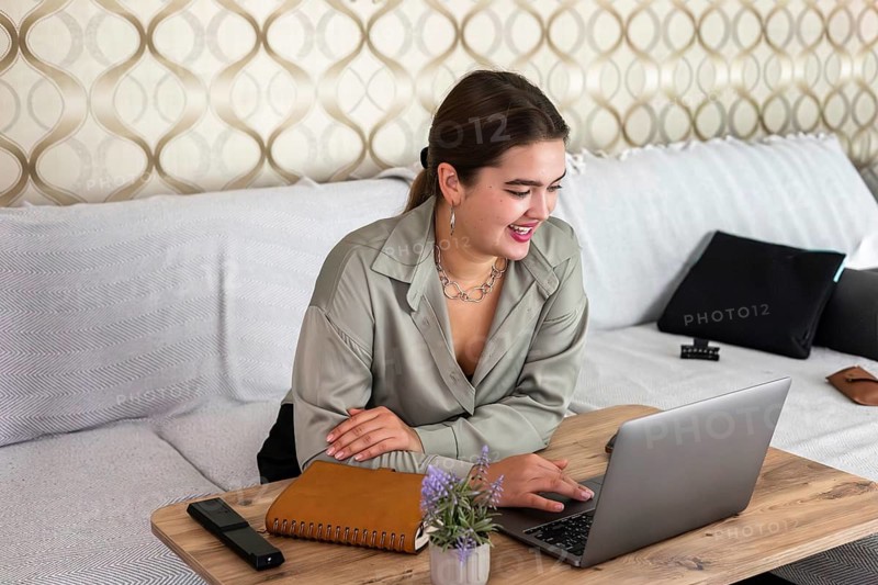 Young Caucasian woman with brown hair working on a laptop at home. She is smiling and sitting at a wooden table with a notebook and a plant