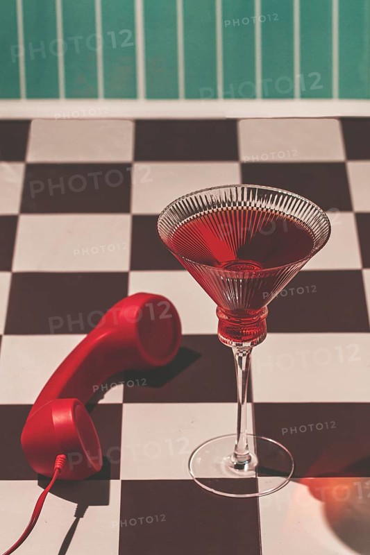 A red drink is served in a clear glass on a black and white checkerboard table. A red phone is next to the drink in a room with green walls