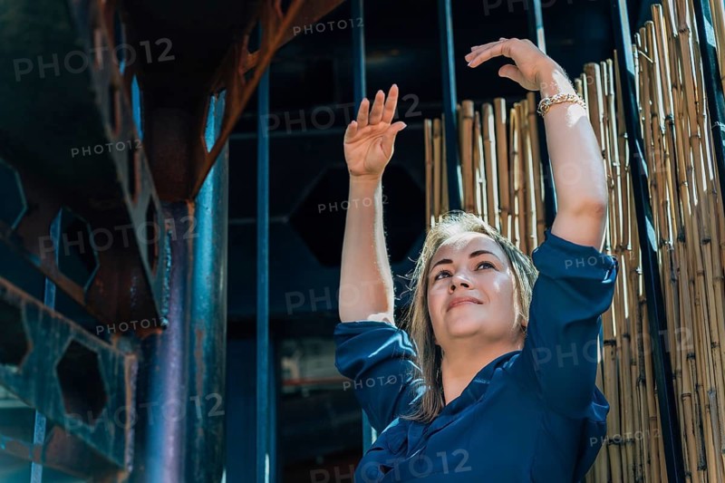 Woman in blue shirt raising arms against a bamboo fence background, natural light, industrial setting, concept of freedom, Woman posing gracefully against a backdrop of bamboo and metal under strong sunlight