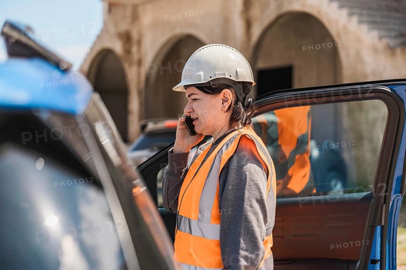 Worker woman in a hard hat and safety vest talks on her phone near a blue car. The scene is set outdoors with a clear sky